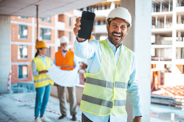 Engineer holding blueprints and showing a smartphone. Male construction engineer demonstrating his...