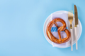 Minimal Oktoberfest table setting. Plate, fork, knife, with festive decorated napkin, checkered ribbon, pretzel, beer glass on blue background. Oktoberfest festival invitation, menu catering flat lay