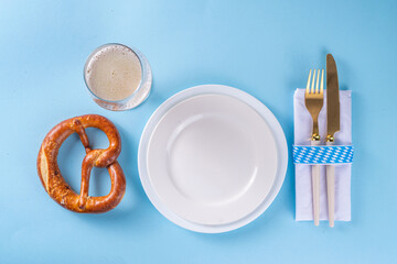 Minimal Oktoberfest table setting. Plate, fork, knife, with festive decorated napkin, checkered ribbon, pretzel, beer glass on blue background. Oktoberfest festival invitation, menu catering flat lay