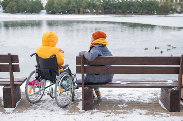 Caucasian woman in a wheelchair and her friend are sitting by the lake with ducks in winter.