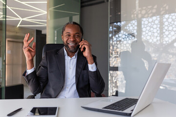 Portrait of successful african american man inside office at workplace, mature businessman talking on phone smiling and looking at camera, satisfied with financial achievement boss with laptop.