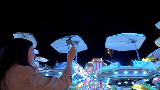 Woman hands are holding smartphone and taking photo or shooting video. Carousel with lights. Incredible colorful flashing light of vintage carousel.Popular Chair Swing Rides in Amusement Park