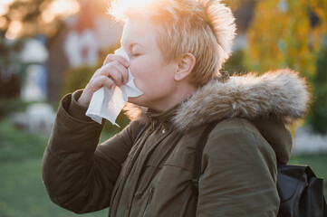 Blonde short cut hair woman in a stylish parka blows her nose into a paper napkin outdoors in an autumn park on yellow leaves background. Seasonal autumn cold