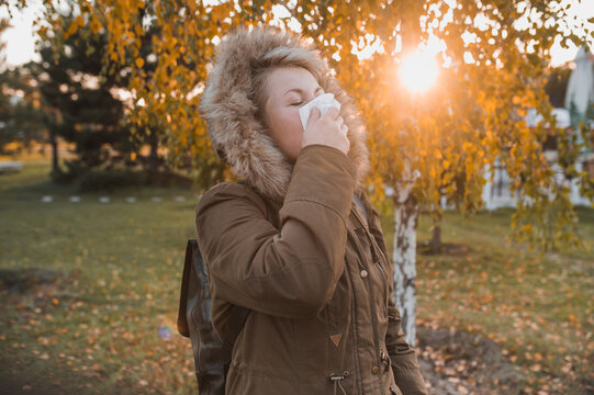 Blonde short cut hair woman in a stylish parka blows her nose into a paper napkin outdoors in an autumn park on yellow leaves background. Seasonal autumn cold