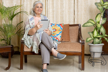  Beautiful Indian senior woman using tablet and smiling while relaxing on the couch at home