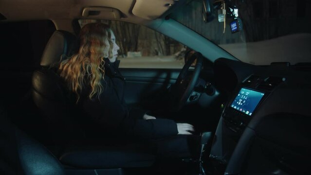 A tired asleep young woman with long curly hair and in a black jacket turns off the engine from the button, unfastens the belt and gets out of the car, closes the door and leaves at night in winter