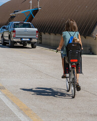 Obraz premium An authentic mother, in a sunny summer morning, carries her daughter in a child seat on a bicycle's trunk, casting a cyclist's shadow on the asphalt road, with a parked car in the background
