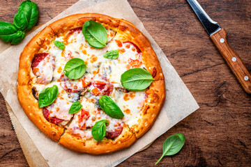 Pizza with italian salami sausage, mozzarella cheese, tomato sauce and green basil on rustic wooden kitchen table background, top view
