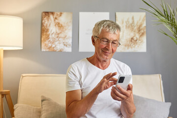 Pleased smiling senior man wearing white T-shirt texting on smartphone scrolling reading news in social media and browsing internet typing sms chatting online.