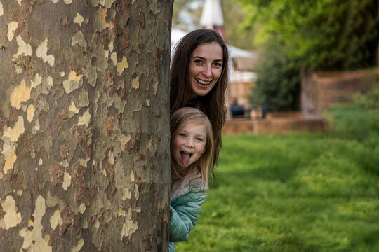 Smiling Mom And Little Blonde Girl Child Peeking Out From Behind A Tree In The Park