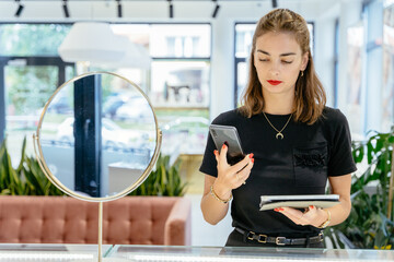 Portrait of a female entrepreneur standing in a jewelry store behind the counter with a smartphone and tablet managing the business, making a call, write message. Luxury jewelry store concept.