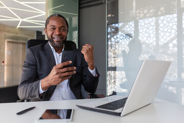 Portrait of successful winner at workplace inside office, African American man smiling and looking at camera, businessman holding phone, received online notification of winning message.