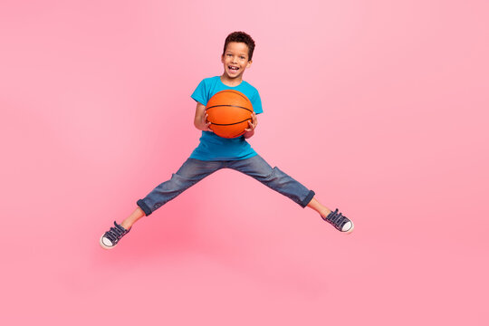 Full length photo of positive impressed small boy wear blue t-shirt playing basketball jumping high isolated pink color background