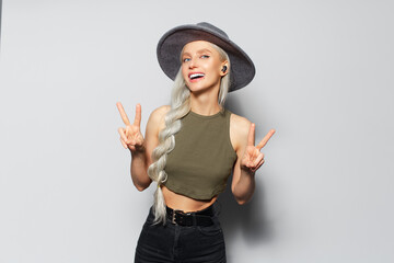 Studio portrait of attractive happy blonde girl with wireless earbuds in ears, showing peace sign, on white background. Wearing grey hat and green shirt.