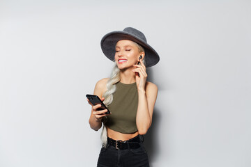 Studio portrait of beautiful happy girl with blonde hair, wearing hat, using wireless earphones and smartphone on white background.