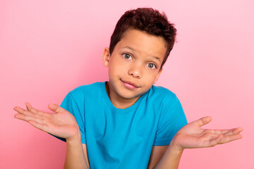 Photo of doubtful unsure small boy wear blue t-shirt shrugging shoulders isolated pink color background