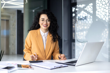 Portrait of mature successful female financier inside office at work place inside office, Hispanic smiling and looking at camera calculating financial figures and checking reports with calculator.