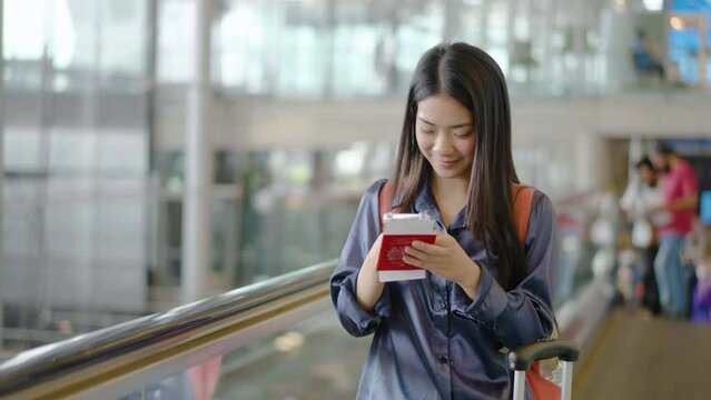Young asian woman checking phone on escalator in airport terminal. Female going up with luggage during travel trip. Girl chatting on smartphone, smiling and waiting for flight. Concept of travel