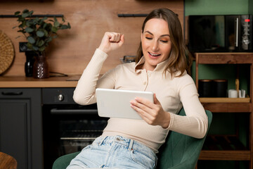 An emotional brunette woman participates in an online auction using a tablet at home, raises her...