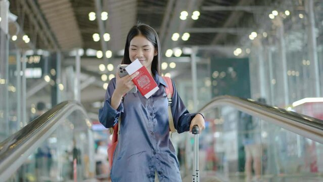 Young asian woman checking phone on escalator in airport terminal. Young woman going down with luggage during business trip, smiling and waiting for flight. Concept of travel.