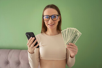 Happy, beautiful young woman with glasses, standing in an excited state, holding an online smartphone and dollar bills in her hands. 
