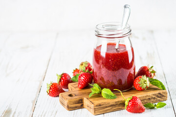 Strawberry jam in the glass jar at white table.