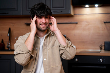 A young man suffering from a severe headache or migraine is sitting in the kitchen, feeling pain concerning the aching head. 