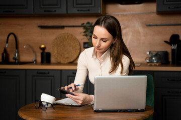 A young brunette woman makes notes makes calculations in a calculator using a laptop in the kitchen. 