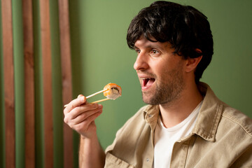 Fototapeta premium Young handsome man wearing beige shirt eating sushi roll with salmon using chopsticks. 