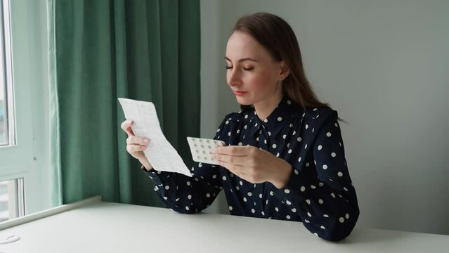 A young brunette woman is sitting at a table in her living room and reading an information leaflet with prescribed medication. 