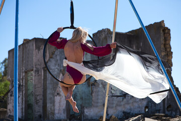 Young and beautiful blonde woman, spectacular exercise on an aerial hoop. She does different artistic and acrobatic poses with a large canvas. The young athlete is in the ruins of a building
