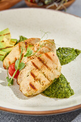 Macro vertical side view capturing the textures of grilled chicken fillet, avocado, and spinach on a ceramic plate. Set against a grey concrete background with hard shadows