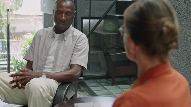 Tilt Down Over The Shoulder Shot Of African American Man Describing Mental Health Problems And Answering Questions Of Female Psychologist As She Making Notes During Consultation