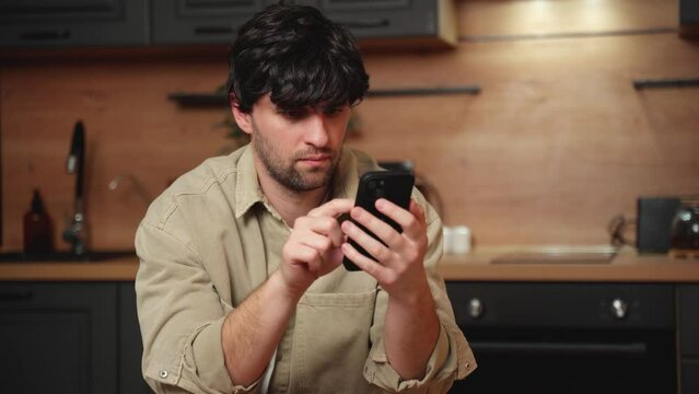 A young brunette man dressed in a beige shirt will scroll his tape on social networks using a smartphone while sitting in the kitchen. 