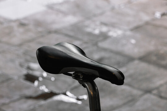 A Foam Cushioning Bicycle Saddle, Or Seat, Without Center Cut Out Shot From A Side Angled View On A Driveway Rider Standing Behind In The Distance, Wheel And Spokes Visible. 