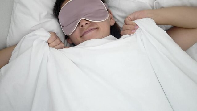 woman with sleeping mask on eyes sleeps on soft pillow joyfully pulling blanket in bed during healthy sleep 
