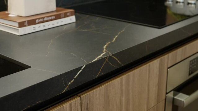 Marble matte kitchen counter with built-in sink, electrical oven and books. Brown wooden cabinets with drawers and integrated stove. Golden pattern on black working surface