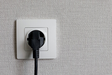 Black power cord cable plugged into european wall outlet on white plaster wall with copy space.