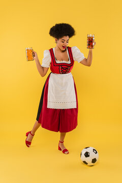 joyous african american oktoberfest waitress holding beer mugs and playing football on yellow