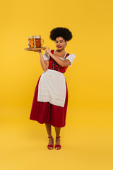 smiling african american oktoberfest waitress holding wooden tray with beer mugs on yellow, banner
