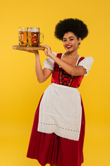 joyful african american bavarian waitress in dirndl serving beer mugs on wooden tray on yellow