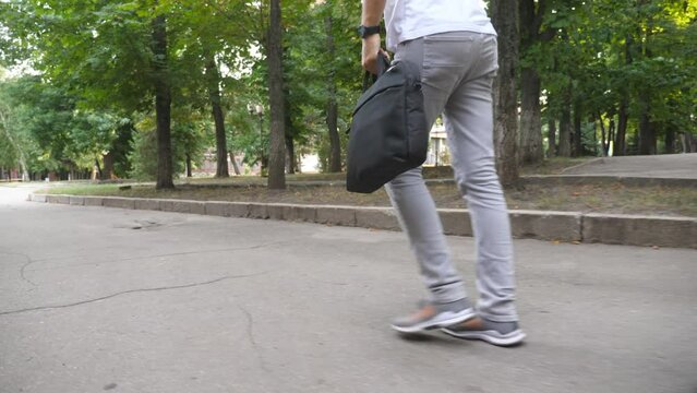 Close up legs of young man in casual wear going through urban park. Businessman with bag looks to his watch and starts running because late to work. Guy talking phone and hurrying at office. Slow mo