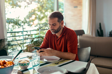 Handsome concentrated man using smartphone while sitting before lunch at home.