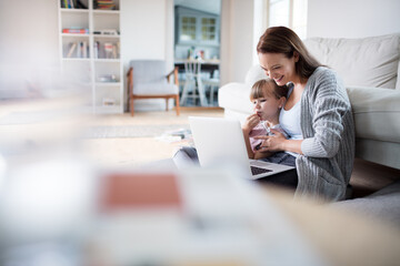 Young mother playing with her daughter while using the laptop in the living room at home