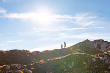 Two silhouettes, hikers walking under the blue sky with white plane trails, contrails, and sun....