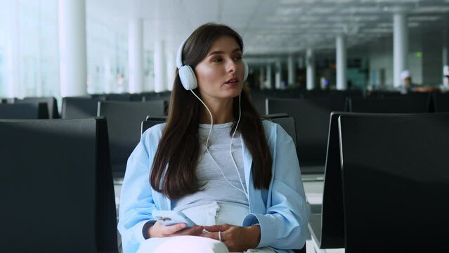 Young woman chilling, listening favorite music via headphones, waiting airplane or train in waiting room with smartphone in hands. Girl waiting boarding at airport. Recreation and vacation concept.