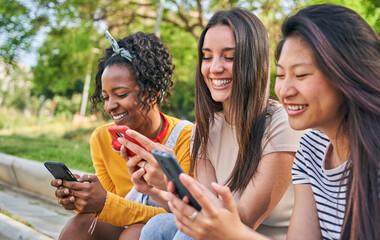 Three smiling young women using cell phone outdoors. Group of happy friends checking fun social networks on mobile. Cheerful classmates gossip on dating apps together. People and new technologies.