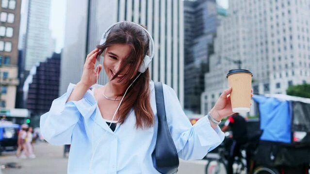 Tourist in new modern big city. Close-up of young pretty woman in headphones listening favorite music, chilling, drinking tea or coffee from glass on warm summer day, looking around cheerfully.