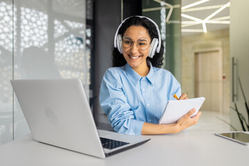Businesswoman inside office watching online video webinar course, Hispanic woman in headphones recording data at workplace smiling satisfied with result of professional development and distance