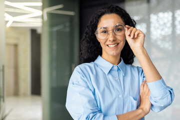 Fototapeta premium Portrait of happy and successful business woman, boss in shirt smiling and looking at camera inside office with crossed arms, Hispanic woman with curly hair in corridor.